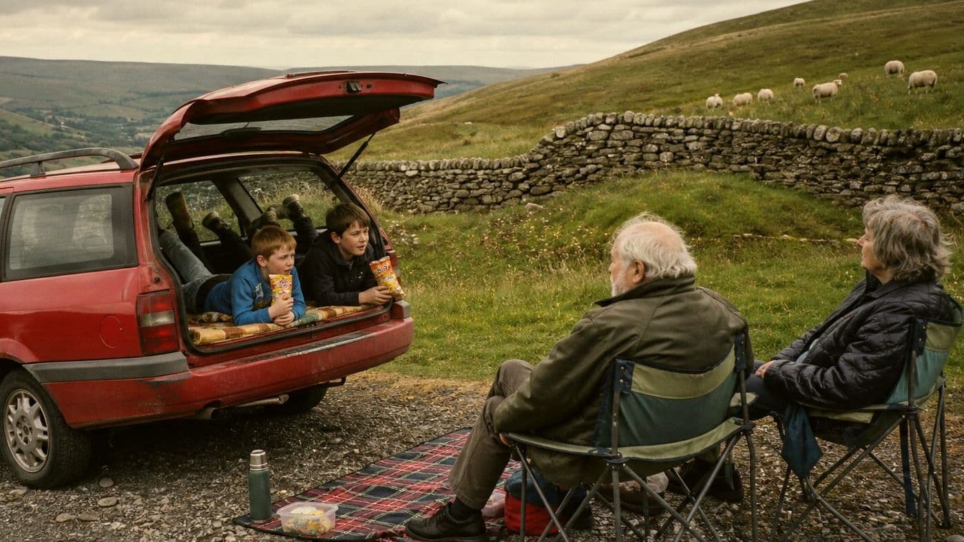 Grandparents in camping chairs beside a red estate car, their grandchildren snacking in its open boot, on a Dales hillside